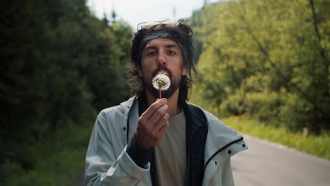 a happy guy tourist in special clothes for hiking blows on a dandelion that scatter against the background of the forest