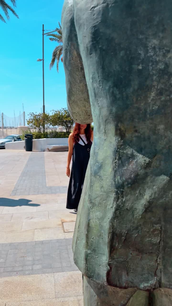 Cartagena, Spagna, 8 agosto 2025: Donna che passa davanti a una statua di bronzo sulla passeggiata del porto di Cartagena. Vista posteriore di una donna dai capelli rossi che cammina lungo un molo moderno vicino a una statua di bronzo e una tettoia bianca