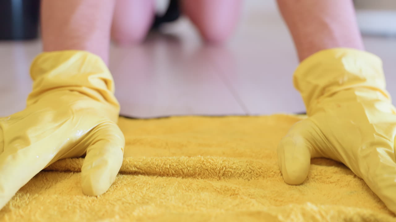 Close up of person wearing yellow gloves moving rag forward and backward on wooden floor, showing household cleaning service, sanitation process, hygiene effort and domestic maintenance