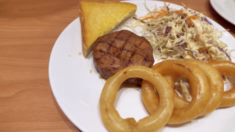 close up of a plate filled with onions rings, toasted bread, coleslaw salad, and a cut of medium rare burger steak, served in a restaurant in bangkok, thailand