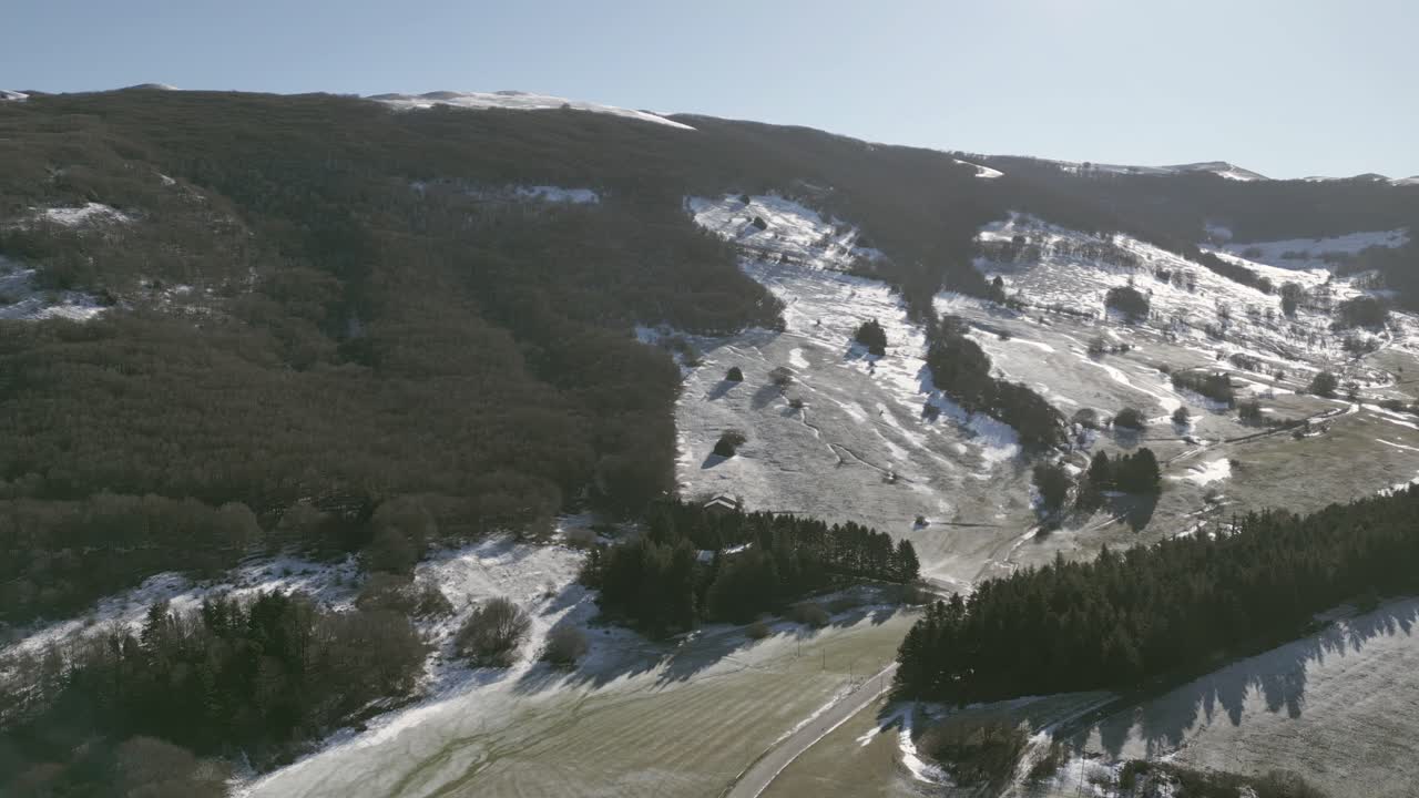 Drone view over Valsorda, Gualdo Tadino, on a sunny winter day. Snow-dusted peaks rise above a vibrant landscape, blending crisp white and rich earthy tones under a clear blue sky
