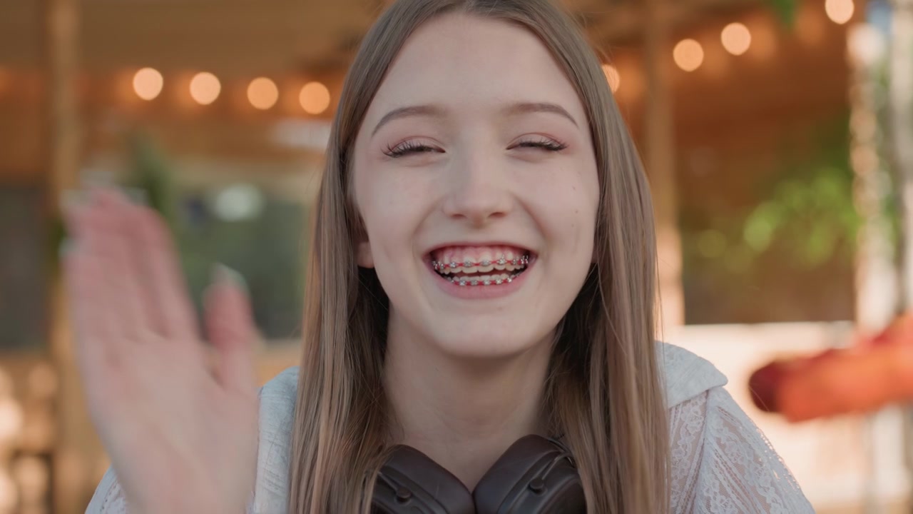 Primo piano di una giovane donna che parla e gesticola con le mani in un ambiente esterno. La donna indossa un top di pizzo di colore chiaro, è visibile un accessorio scuro al collo, capelli lunghi e lisci. Lo sfondo presenta un filo.