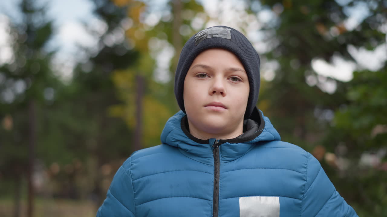niño con gorra negra y chaqueta azul de pie al aire libre, ajustando y cerrando su chaqueta en un día fresco de otoño, rodeado de árboles borrosos y naturaleza