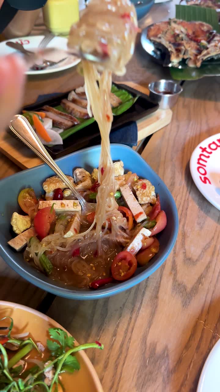 Tangan Mengaduk dan Mengangkat Mi Kaca Yum Woon Sen dalam Suasana Makan Santai dan Cerah di Bangkok
