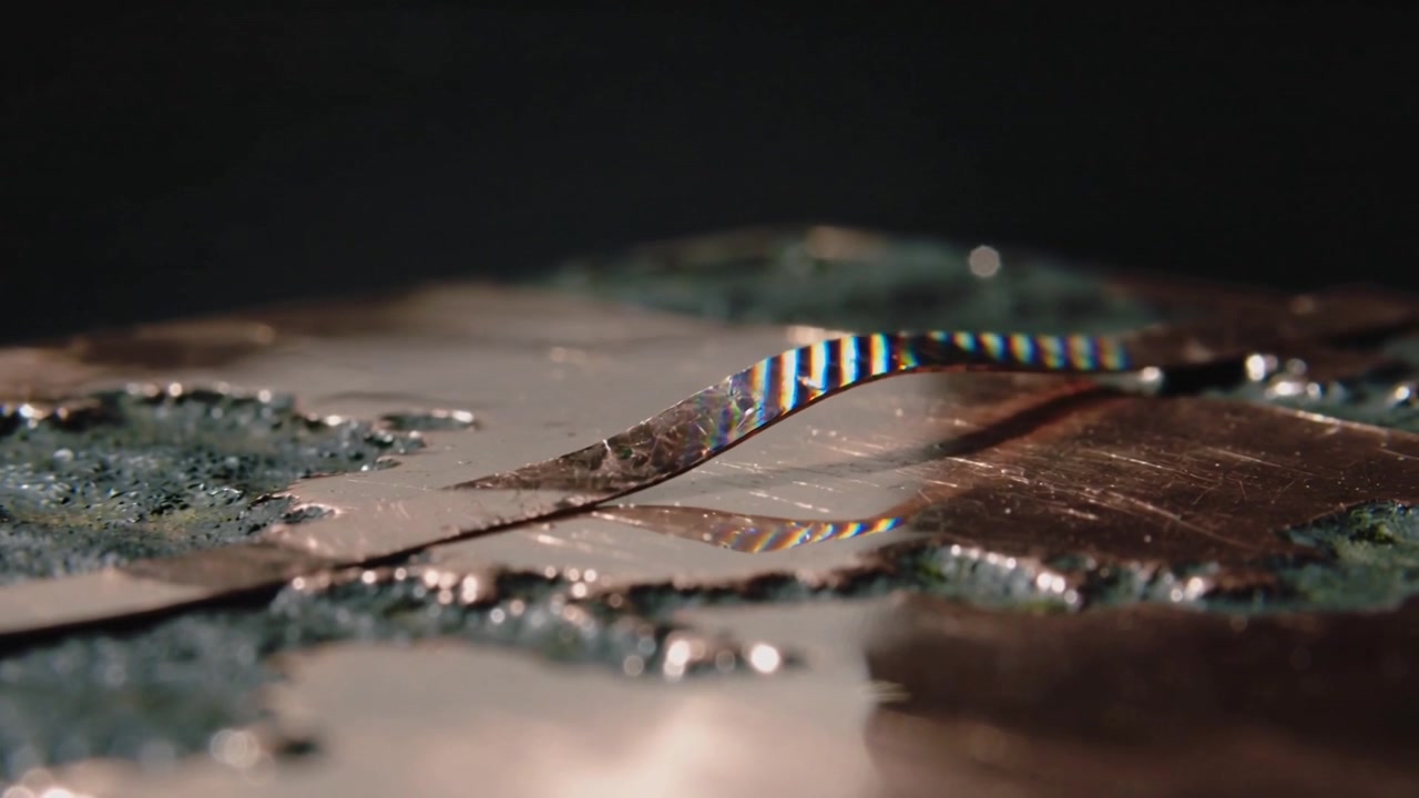 Shifting light lifting foil ribbon into arced loop on pitted copper plate, showing iridescent bands