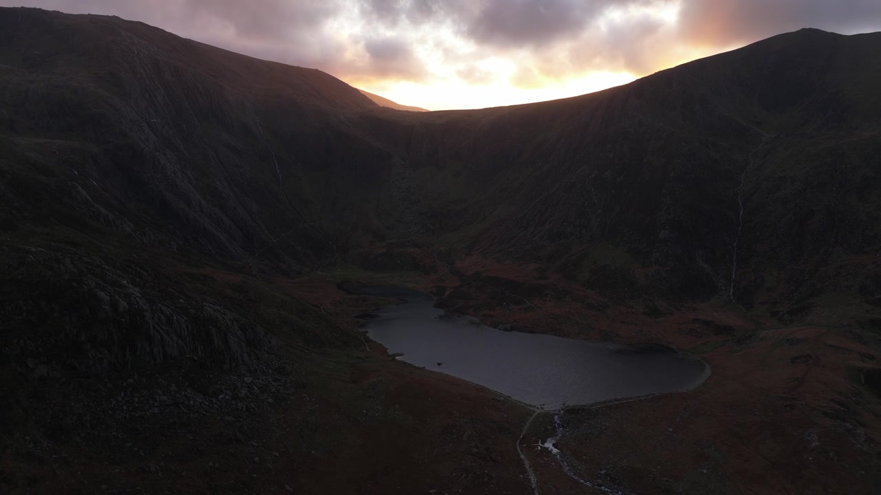 Dramatic golden light from the setting sun moving over a moody mountain valley, illuminating the clouds and reflecting on a remote alpine lake, capturing a serene and wild landscape at dusk