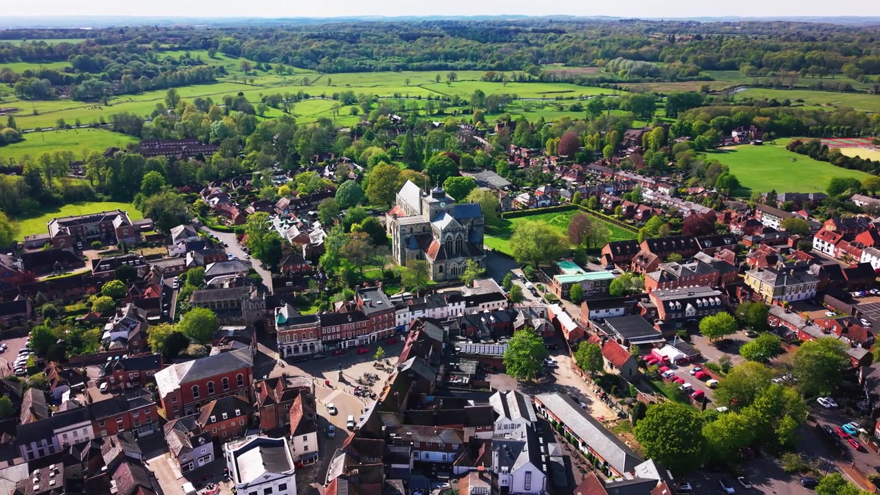 Riprese aeree con drone ad alta quota dell'Abbazia di Romsey nell'Hampshire, che mostrano la storica chiesa abbaziale, il centro città, i tetti, le strade, gli spazi verdi, gli alberi e la campagna circostante in una giornata di sole.