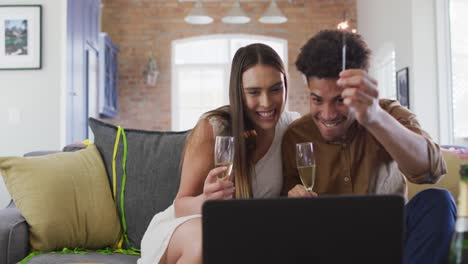 Happy-biracial-couple-drinking-champagne-at-home-making-celebration-laptop-video-call-and-laughing