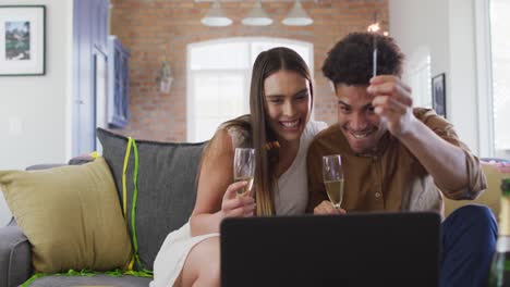 feliz pareja biracial bebiendo champán en casa haciendo una celebración llamada de video portátil y riendo