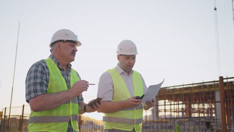 waist-up of two middle-aged male builders wearing safety clothing standing at construction site man using walkie-talkie his colleague holding paper with project plan