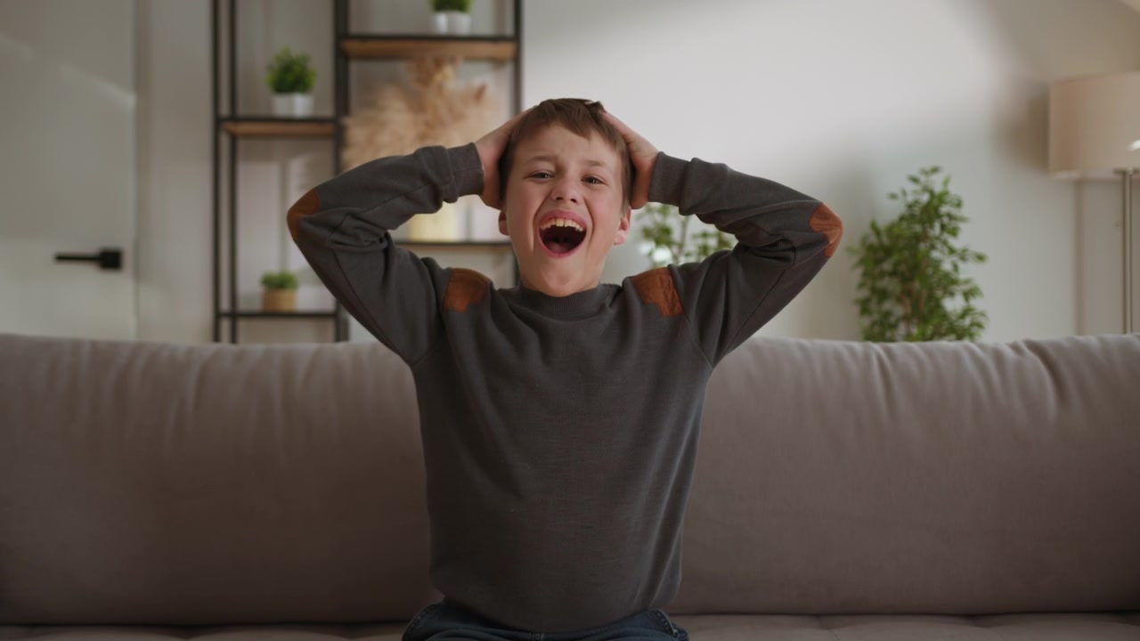 Young boy sitting on sofa hands on head screaming in frustration and distress overwhelmed by emotion