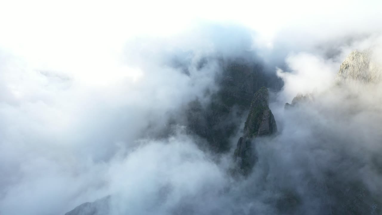 Drone shot circling around the foggy and cloudy peaks of Pico das Torres in Madeira as the morning sun peaks through