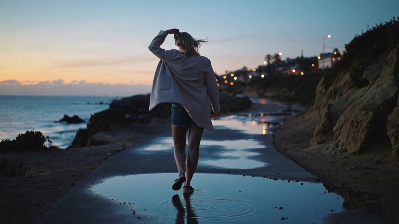 Walking woman in light coat heading toward lit houses along coastal path at dusk, stepping puddles