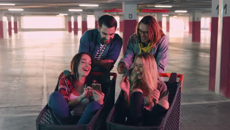 caucasian group of friends watching something on the smartphone and laughing while two of the girls are sitting in a trolley in a empty park