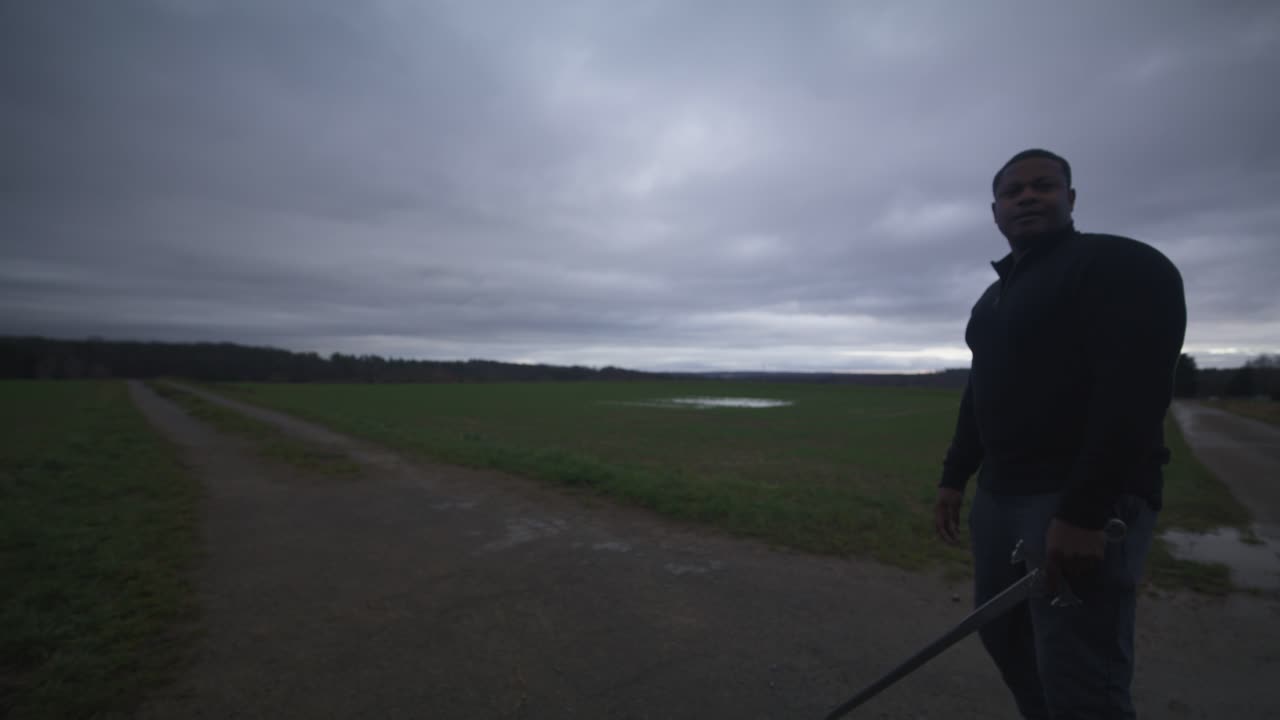 현대의 검은 검사 전사는 회색 하늘 아래 야외에서 대결 상대를 기다리고 있습니다. - 유럽, 4k - 근육, 협박, 그림자, 형상, 흐릿한, 어두운 옷, 강력한, 조정 입장, 심각한.