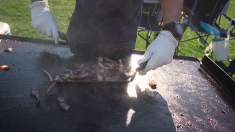 Man-tossing-grilled-onions-with-spatulas-on-hot-grill-in-slow-motion,-close-up-for-backyard-bbq-with-friends