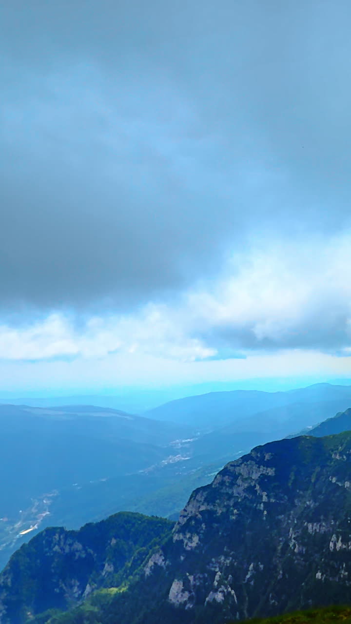 Cima rocciosa verso il fondo di una profonda valle con una strada tortuosa sotto un cielo cupo. Pendii montuosi verdi e valle distante. Video verticale.
