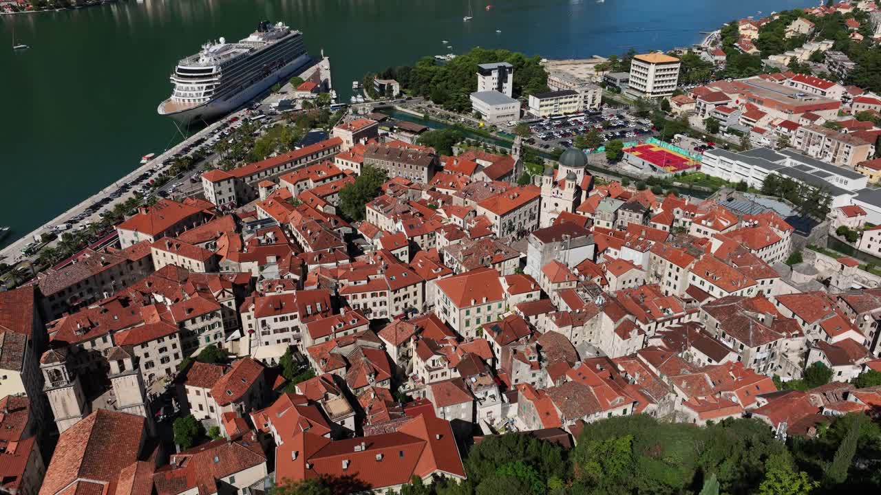 Experience a smooth aerial left panning view across the rooftops of the old walled city of Kotor in Montenegro, revealing the intricate network of buildings and the surrounding landscape