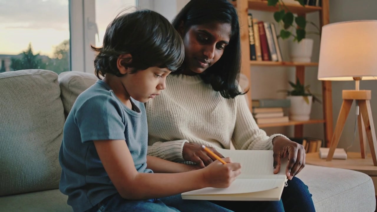Opening notebook kid practicing writing, mother guiding on couch at home with yellow pencil