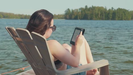 camera pans over a woman relaxing and reading on a dock next to a lake in the summer season