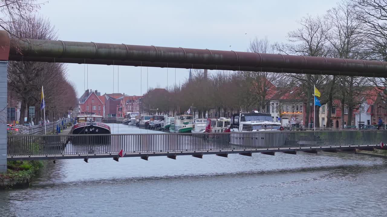 Architectonische voetgangersbrug in Brugge met op de achtergrond een rondvaartboot die over het kanaal vaart, wat het historische stadsbeeld en de waterwegen benadrukt