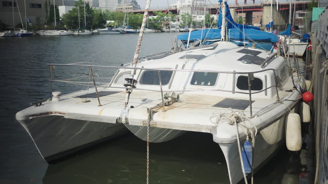 Amidst a dock filled with boats, a worn and weathered white catamaran sailboat finds its place of rest in the vibrant neighborhood of Greenpoint, Brooklyn