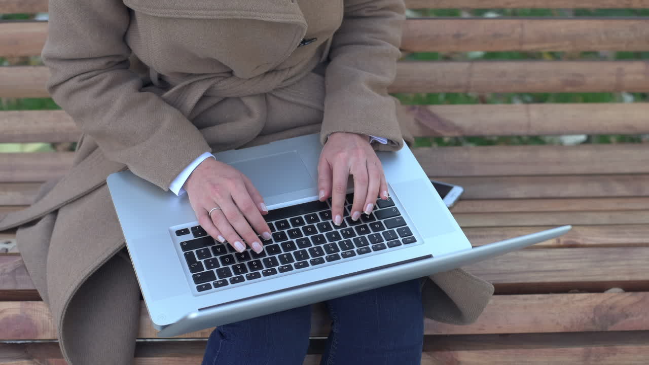 Woman in a brown coat working on her laptop on a bench in the park