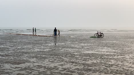 silhouette view of a beach with waves, people taking net for fishing in front of cycle and some people talking in a empty beach in india