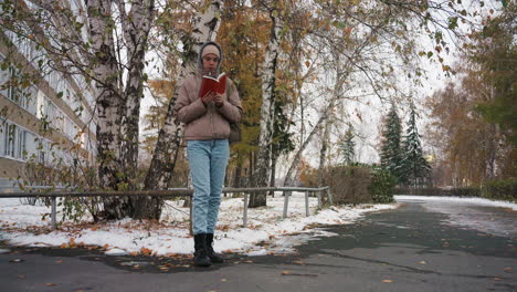light skin lady dressed in winter jacket, hoodie, and beanie stands on snow-covered sidewalk near birch trees, deeply focused reading book during cold autumn day, surrounded by soft natural colors