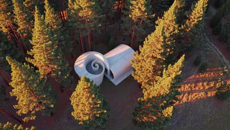 aerial view capturing a unique spiral shaped concrete house nestled in a dense pine forest, glowing warmly in the sunset, highlighting innovative architecture harmonizing with nature