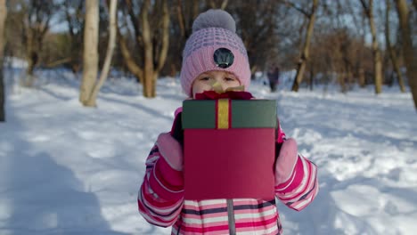 Lächelndes-Kind-Im-Verschneiten-Winterwaldpark,-Das-In-Die-Kamera-Blickt-Und-Eine-Geschenkbox-Mit-Weihnachtsgeschenken-Hält