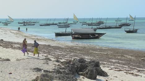 two muslim women walk along a beach in zanzibar with dhow sailboats in the background