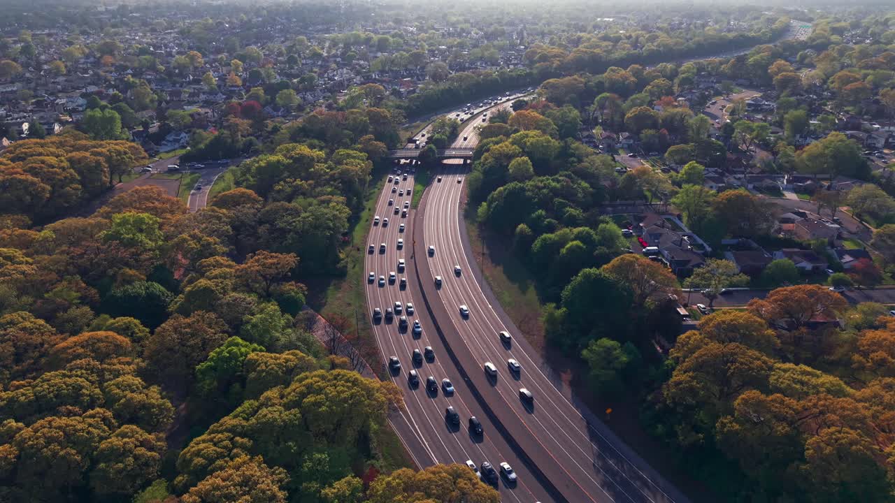 A high angle, aerial view beside the Southern State Parkway on Long Island, NY, taken during a sunny day. The drone hovers by the parkway, looking downward from above green treetops.