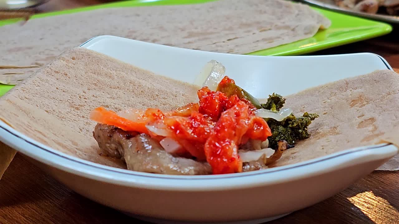 Primo piano estremo di mani che riempiono e arrotolano una crepe di grano saraceno con fette di maiale bollito, kimchi, daikon sottaceto e carote in uno stretto involtino bossam in un ristorante coreano