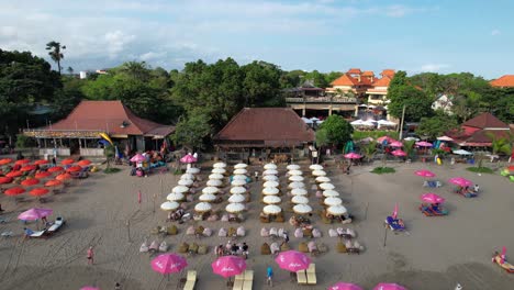 People-Relax-On-Sandy-Double-Six-Beach-Under-Rental-Umbrellas-in-Seminyak,-Bali,-Indonesia---Drone-Flying-Back-Tilting-up