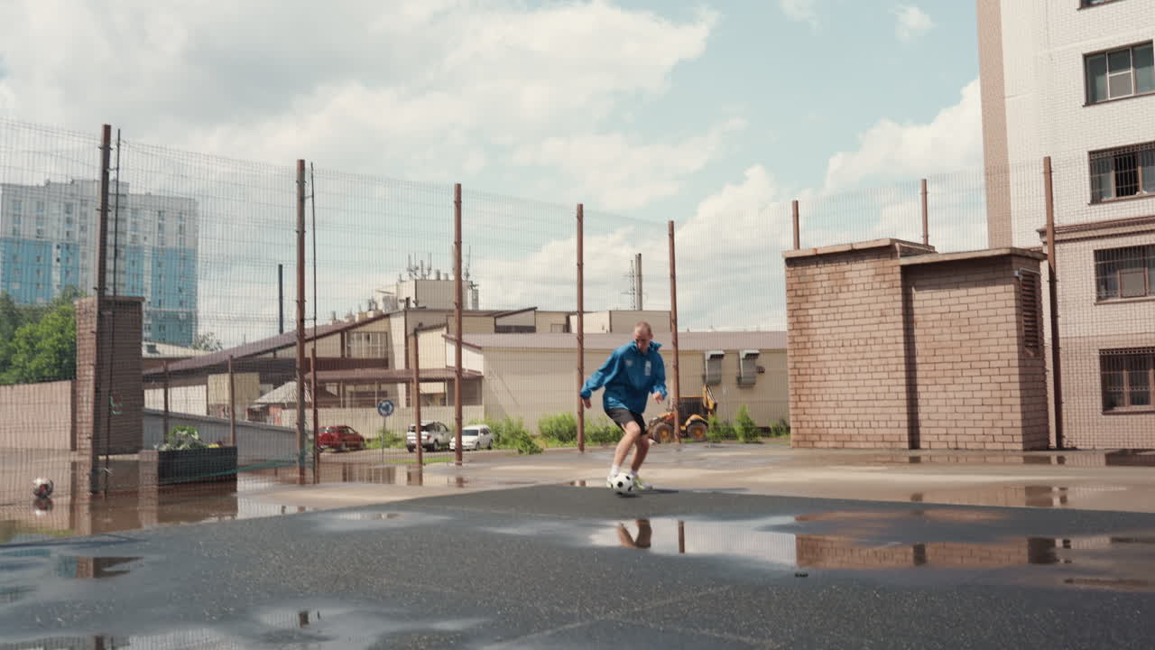 Giocatore di street football che palleggia tra le pozzanghere, giacca blu su campo urbano bagnato, recinzione metallica e palazzi sullo sfondo, primo piano sulla scarpa che colpisce il pallone, spruzzi d'acqua e riflesso