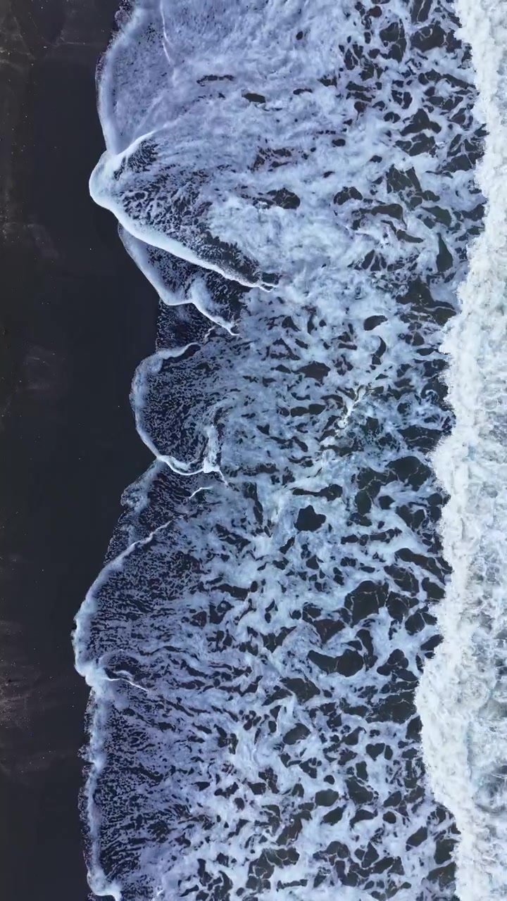 Onde si infrangono sulla sabbia scura della spiaggia di giorno, con schiuma che si forma lungo la riva in una scena costiera.