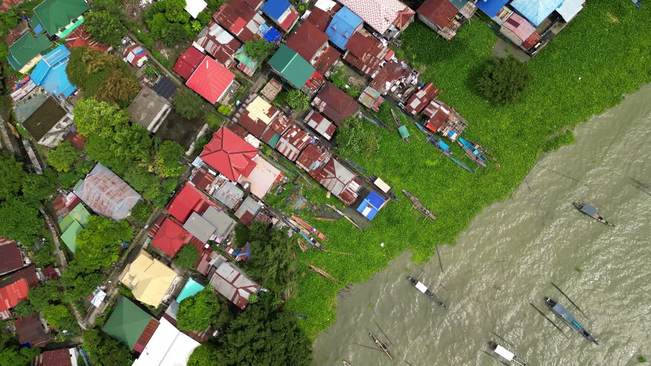 High-altitude drone view captures a dense riverside village with boats, water hyacinths, and rusted rooftops beside forested terrain