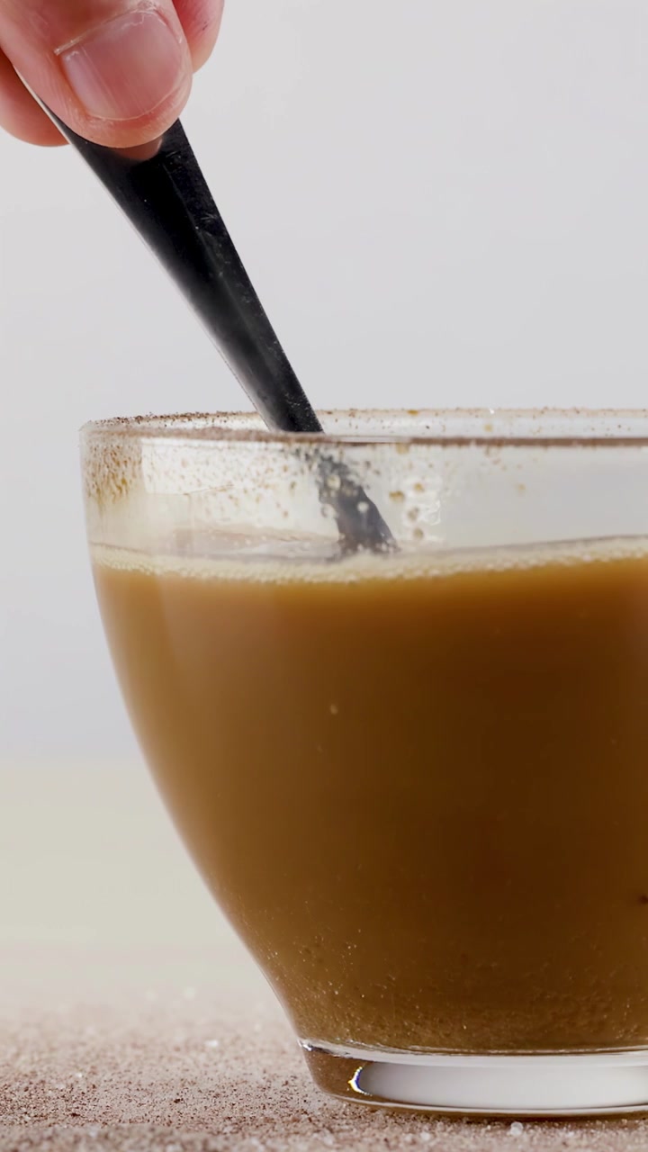 Primo piano di una mano che mescola caffè caldo in una tazza di vetro su sfondo bianco.