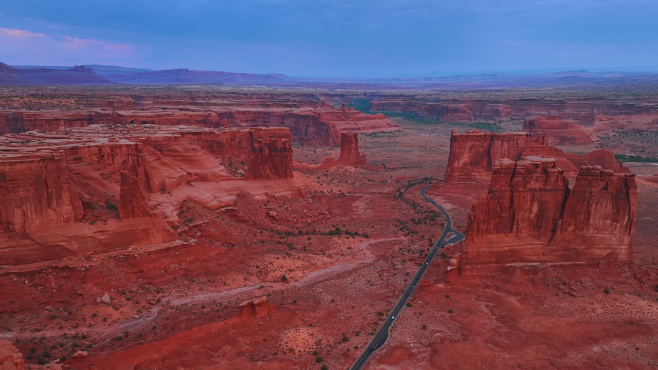 Stort, øde landskap i National Arches Park i Utah, USA, med spektakulære, røde steiner. Noen få biler kjører forbi motorveien som krysser landskapet. Flyfoto