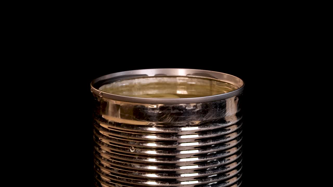 A metal can filled with longan fruit in syrup is struck, causing a dramatic splash. Studio lighting highlights the reflective surface against a black backdrop