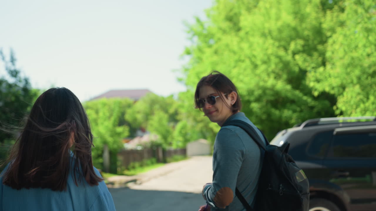 흰색 옷을 입은 남성과 여성이 잎이 무성한 인도에서 백팩을 메고 시선을 교환해요. 길가에는 주차된 차들이 있고, 은은한 여름 햇살이 비치며, 교외 주택가를 지나갈 때 미묘한 사회적 긴장감과 우호적인 유대감이 느껴져요.