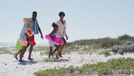 Glückliche-Afroamerikanische-Familie-Mit-Strandausrüstung,-Die-An-Einem-Sonnigen-Tag-Am-Strand-Spaziert