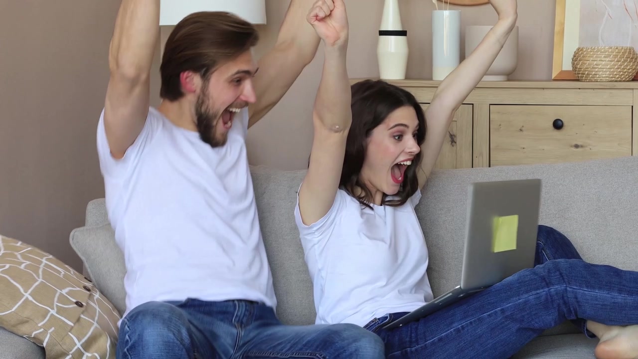 Pareja joven feliz celebrando el éxito mientras miran un portátil en casa