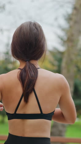 portrait view of sporty woman stretching her back in park with trees in background, focusing on flexibility and stretching exercises for health and fitness, active outdoor workout routine