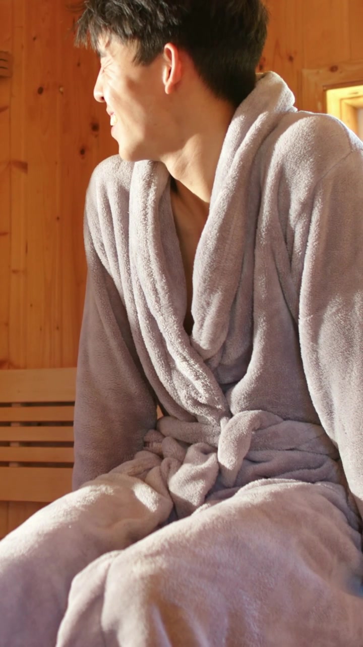Vertical video: Man shifting, turning toward window on bench, sunlight drawing gaze, spa robe