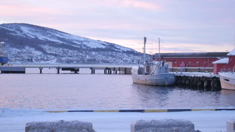 Panning-shot-across-ships-and-boats-moored-at-dock-and-pier-during-polar-night-above-the-arctic-circle-in-Narvik,-northen-Norway-with-snow-covered-hills-in-the-background