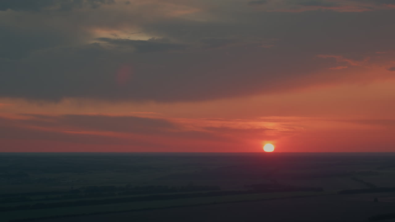 Vibrant red and orange sunset sky with dramatic dark dusk clouds forming shapes while sun glow casts subtle lens flare over expansive flat farmland horizon