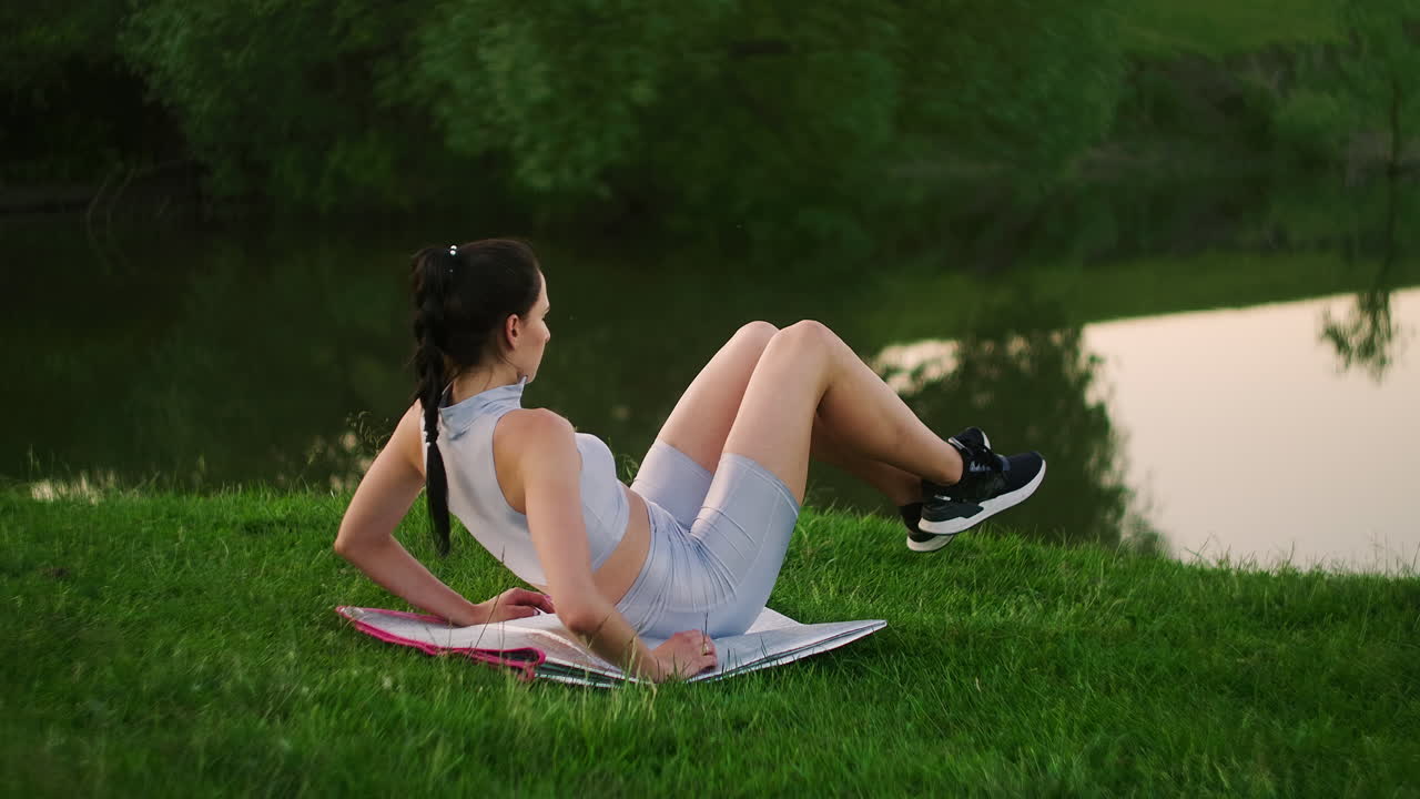 In the morning in the Park a woman in sports clothes lying on a Mat raises her legs to the body. Exercises for a beautiful body. The abdominal muscles