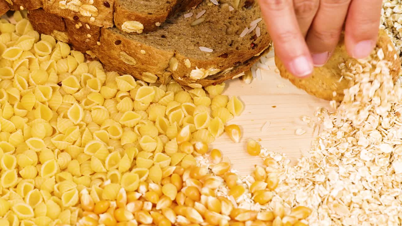 A hand organizes various grains and bread on a wooden surface under bright lighting, creating a visually appealing arrangement